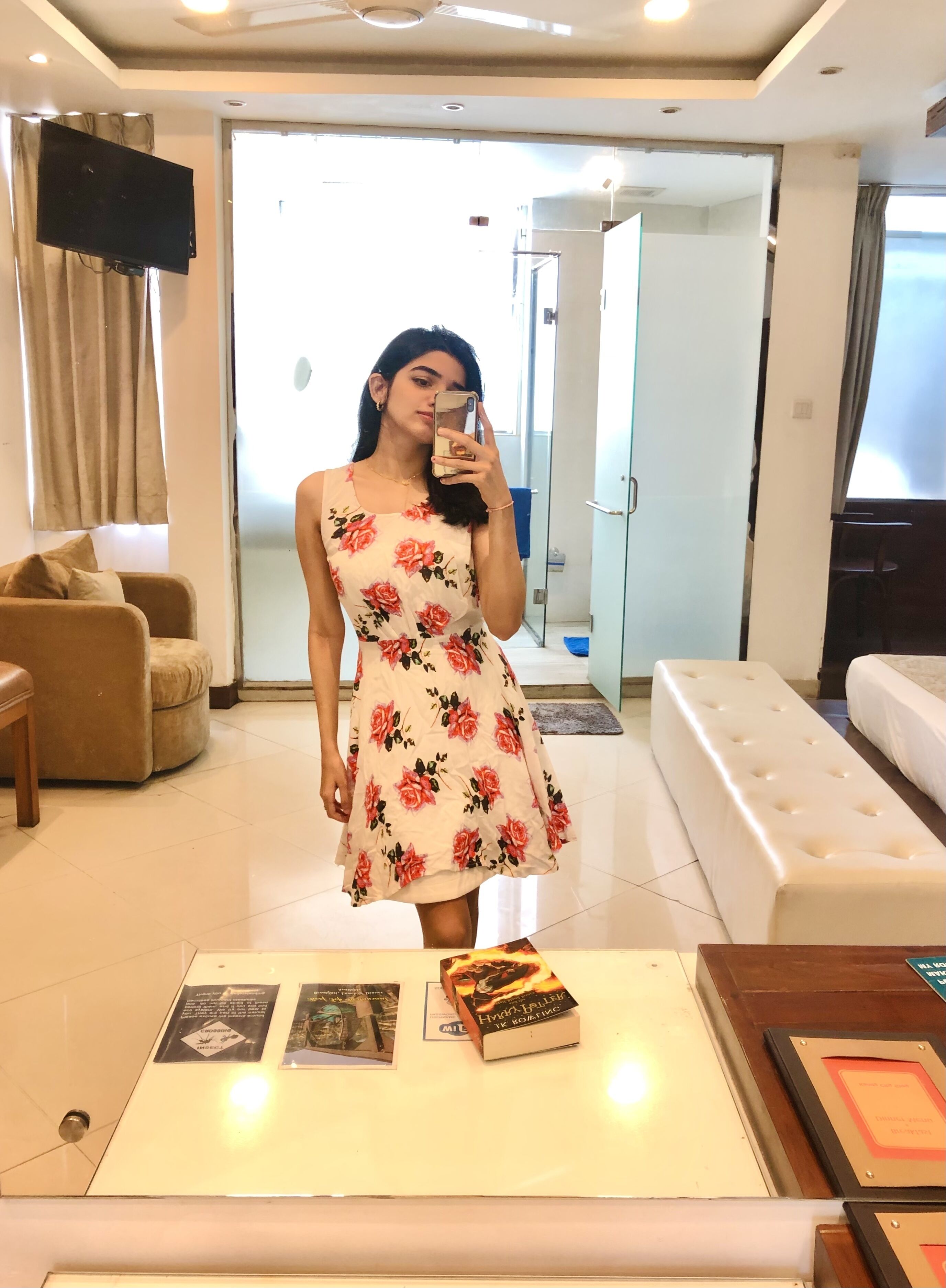 Een vrouw in een bloemenjurk maakt een selfie in een moderne woonkamer met een bank, een bijzettafel met boeken en een televisie.