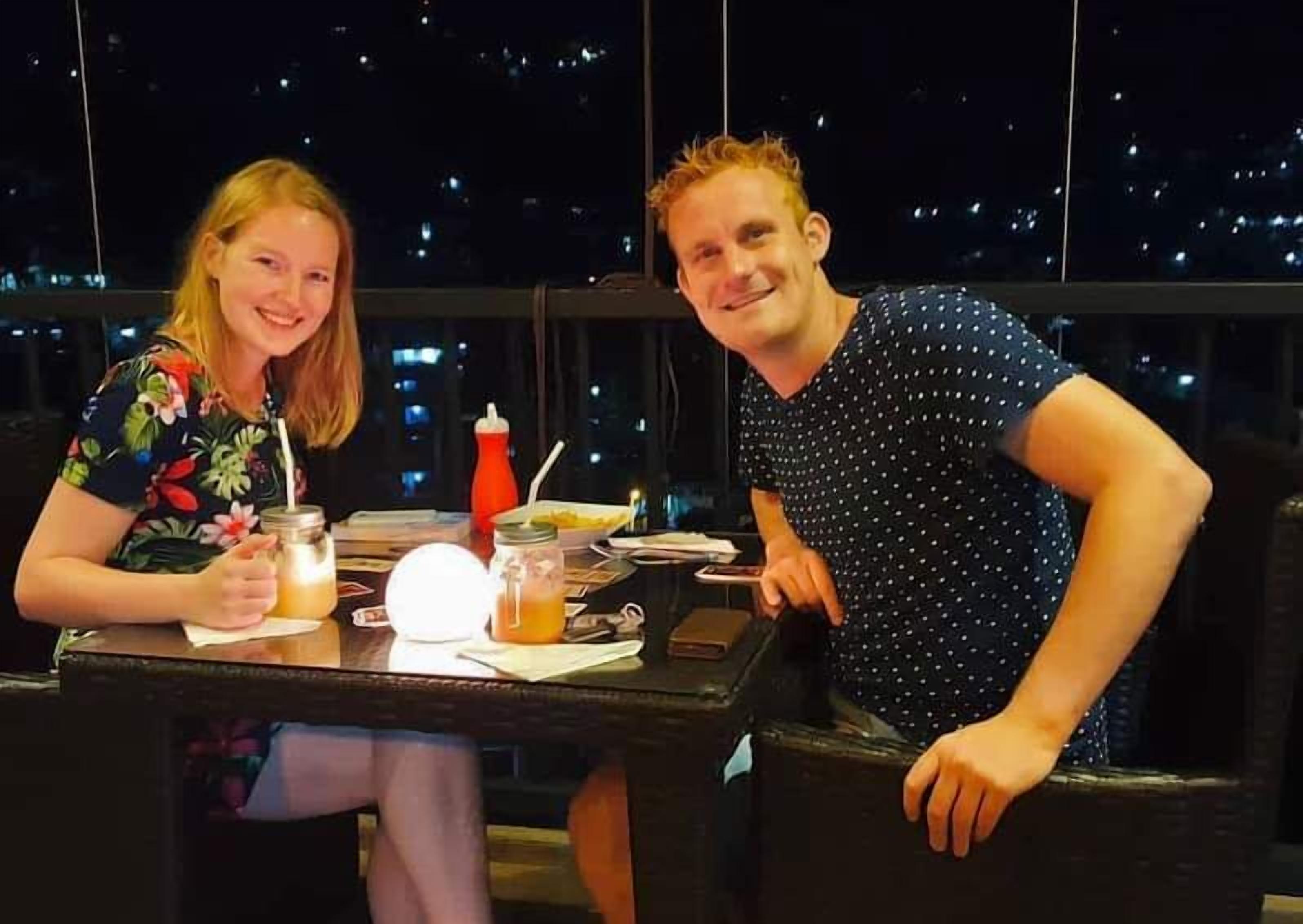 Two people seated at a table with drinks and a bottle of water, in a room with a view of city lights.