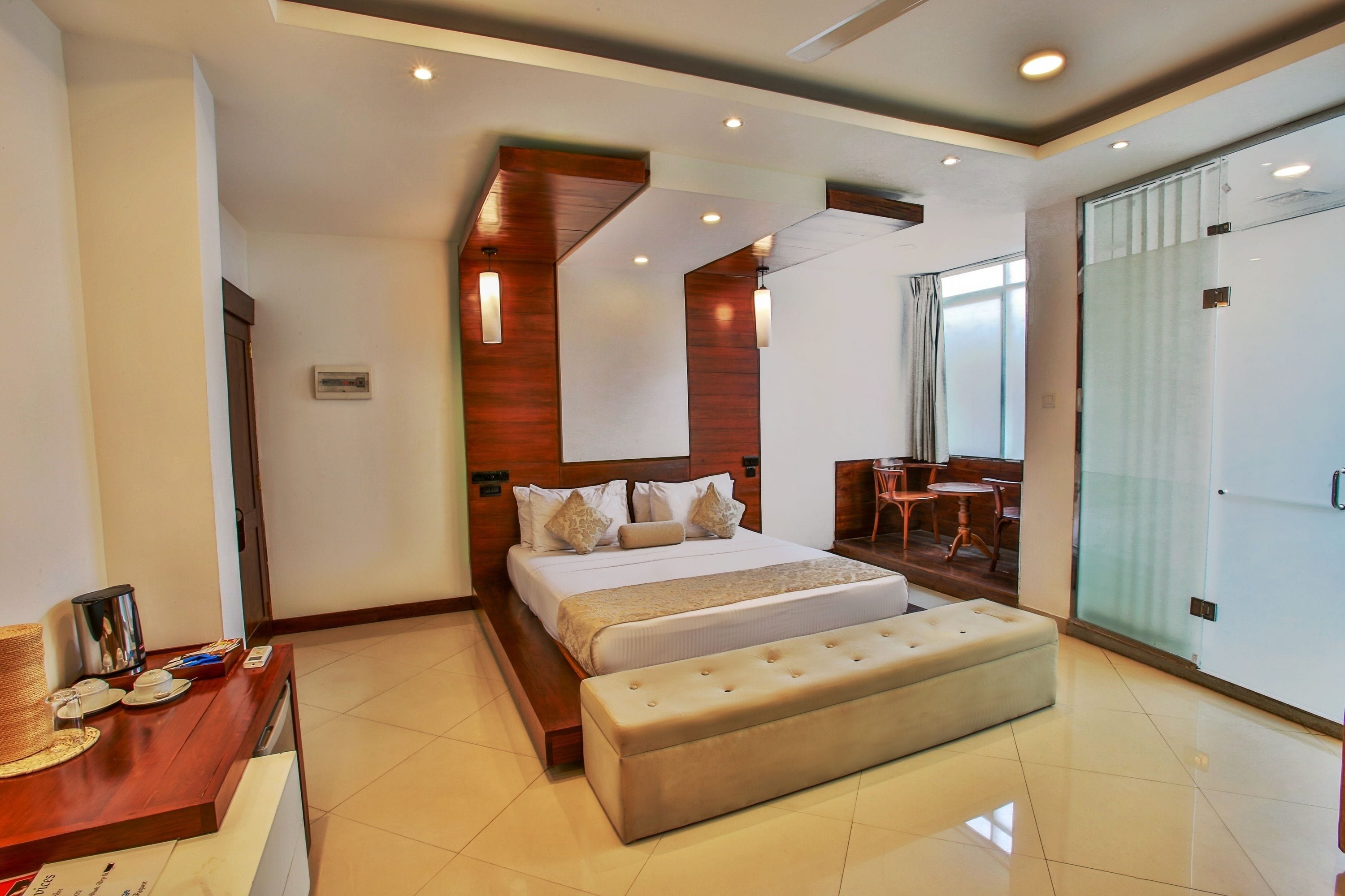 A modern hotel room with a large bed, a wooden headboard, a small seating area, and a glass door leading to a balcony.