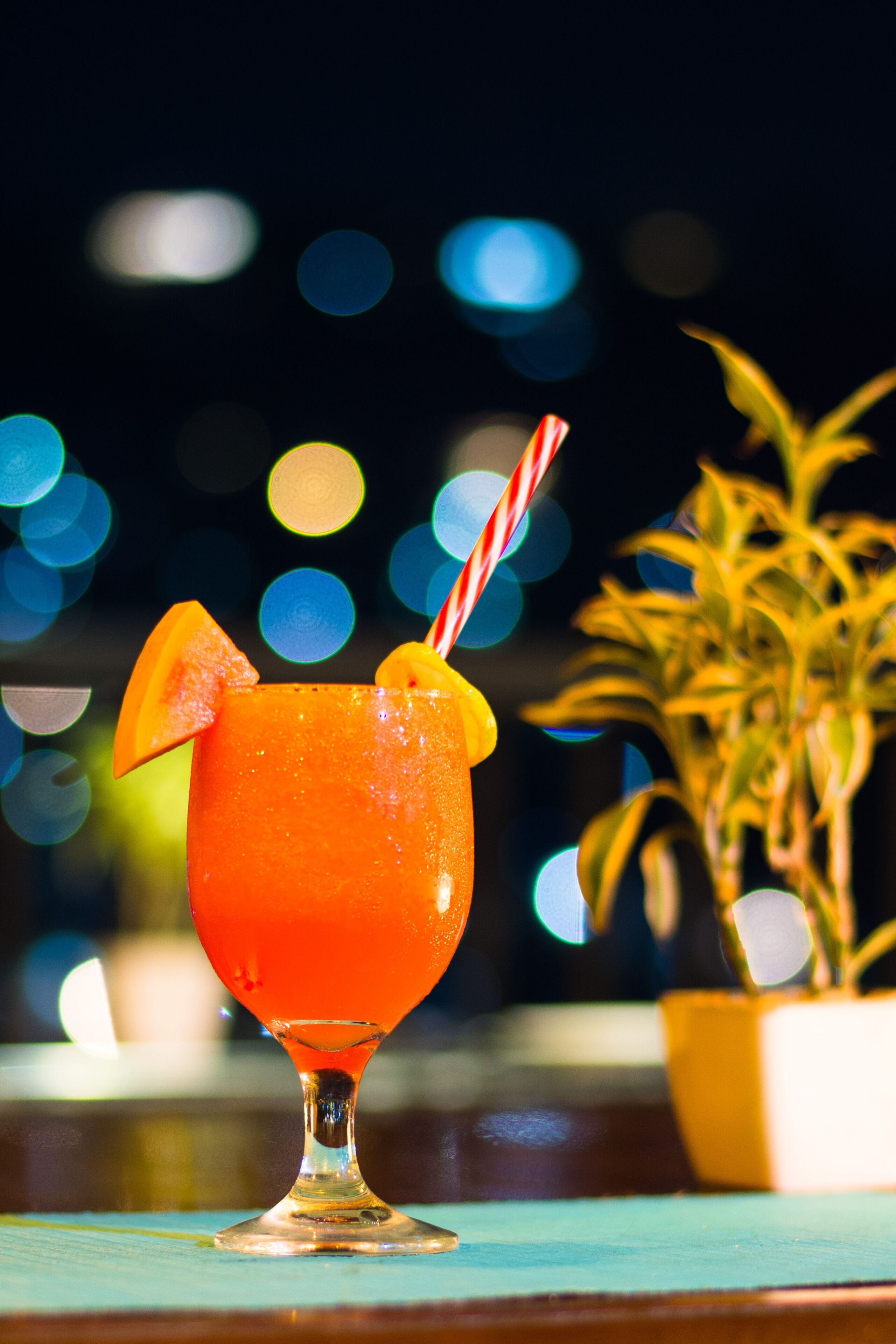 Ein Cocktailglas mit einem gestreiften Strohhalm und einem Orangenscheibchen am Rand, auf einer Holzfläche mit unscharfem Lichthintergrund.