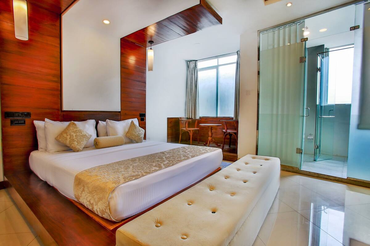 A modern hotel room with a large bed, a bench, a glass shower, and a small table with chairs.
