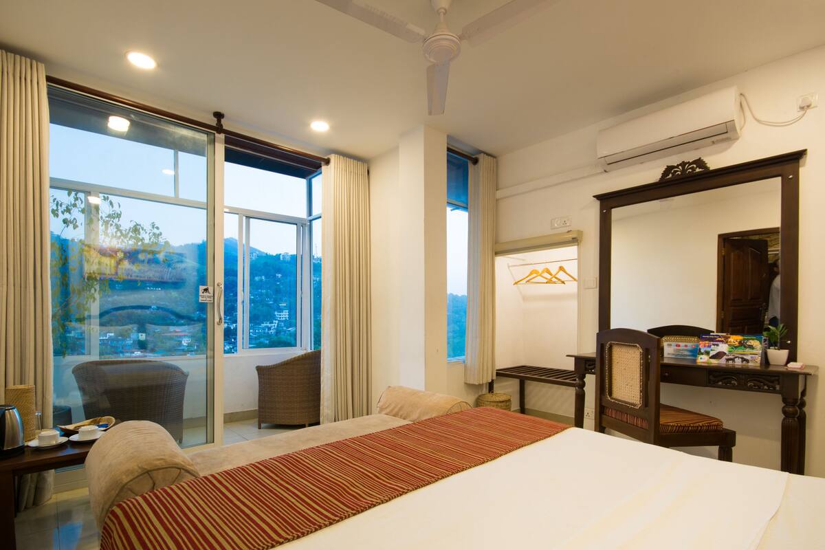 A hotel room with a large bed, a desk with a chair, a window with curtains, and a view of the city.