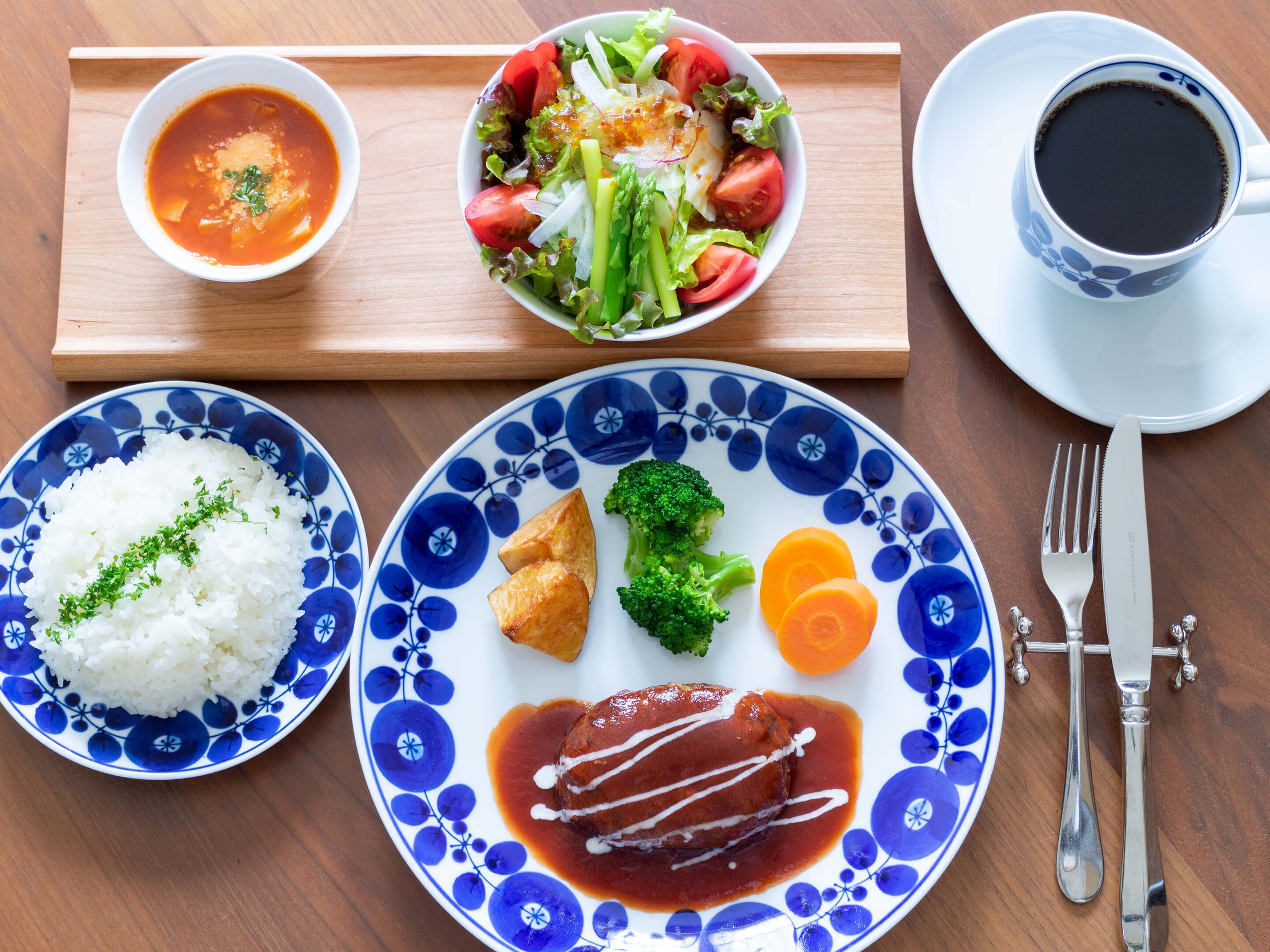 Uma mesa com uma tigela de arroz branco, um prato com carne, uma sopa, uma salada e uma chávena de café.