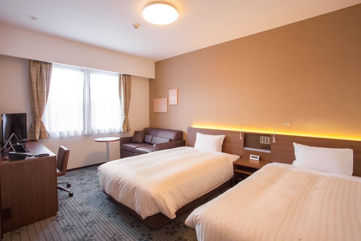 A hotel room with two beds, a desk, a chair, a small table, and a TV.