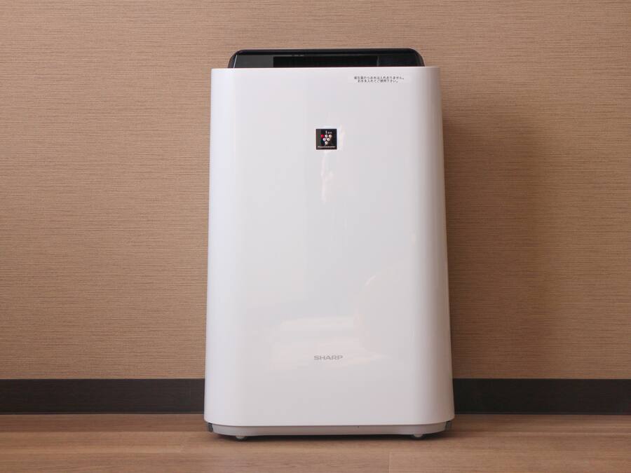 A white air purifier with a control panel on top, standing on a wooden floor against a textured wall.