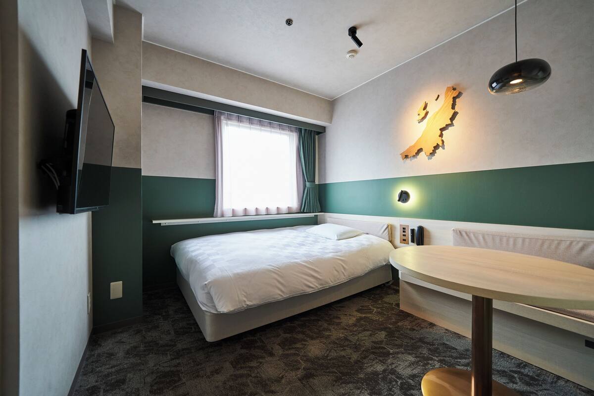 Een hotelkamer met een bed, een bureau, een flatscreen televisie aan de muur, een raam met gordijnen en een wandlamp.