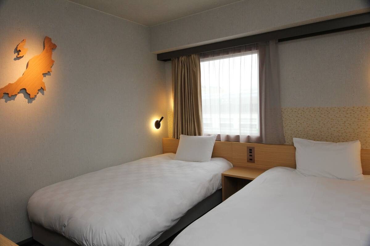 Een hotelkamer met twee eenpersoonsbedden, een houten hoofdbord, een wandlamp en een raam met gordijnen.