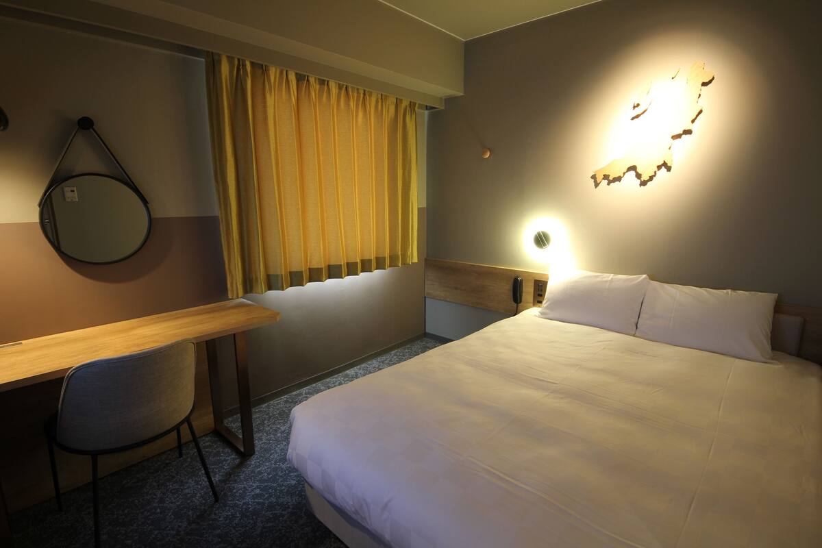 Hotelkamer met een bed, een bureau, een stoel, een raam met gordijnen, een wandlamp en een decoratie van een kaart.