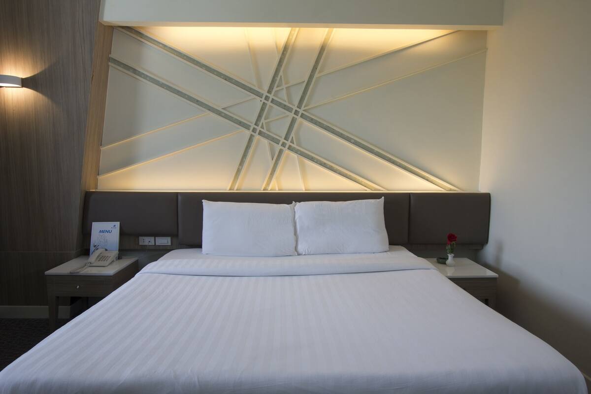 A hotel room with a large bed, a nightstand with a phone, and a wall with a geometric light design.
