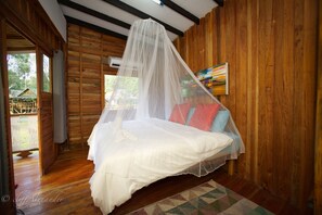 Un dormitorio de cabaña de madera con una cama bajo una mosquitera, un cuadro colorido en la pared y vistas al exterior.
