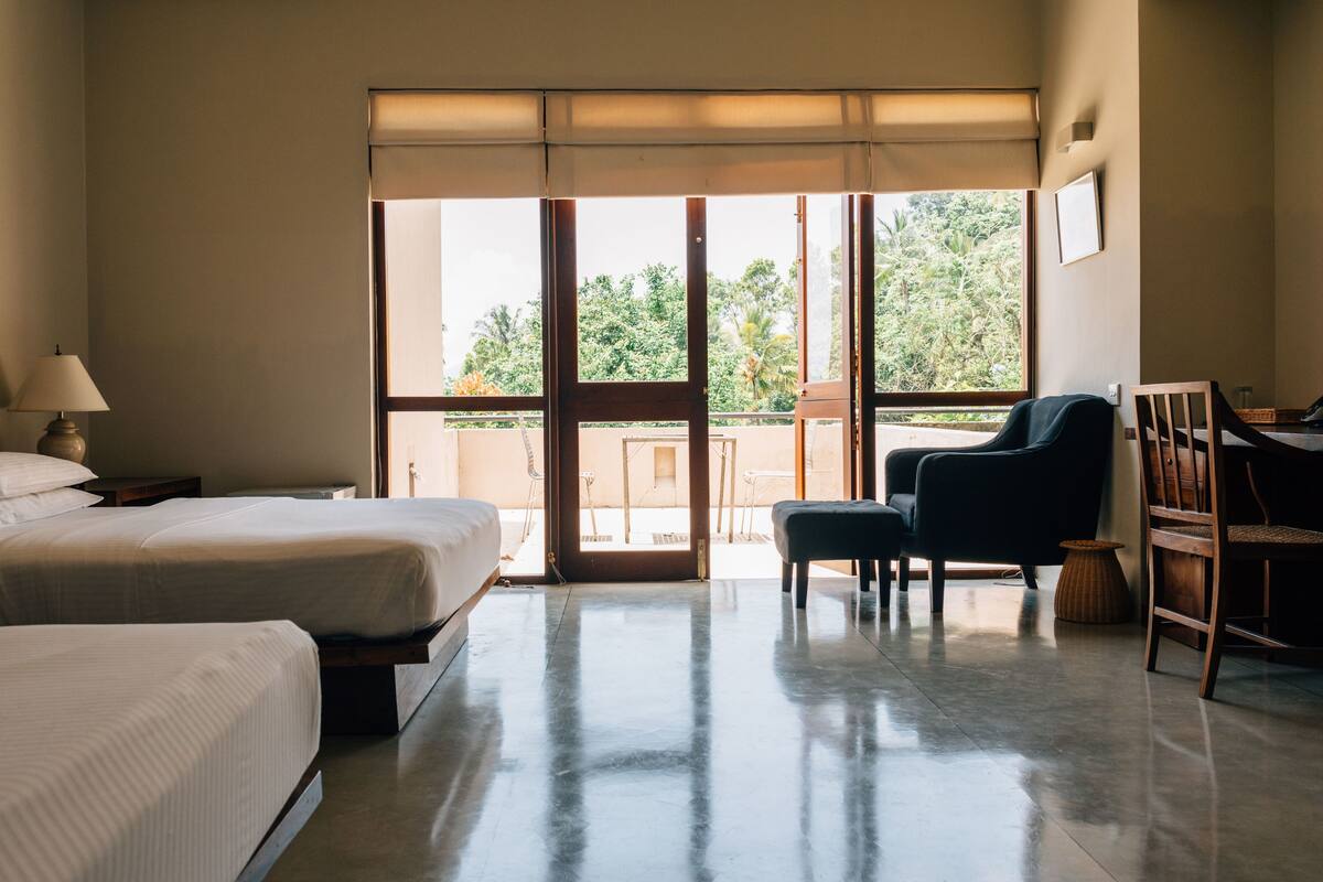 Habitación de hotel con dos camas, una silla, un escritorio y un ventanal con vistas a zonas verdes.