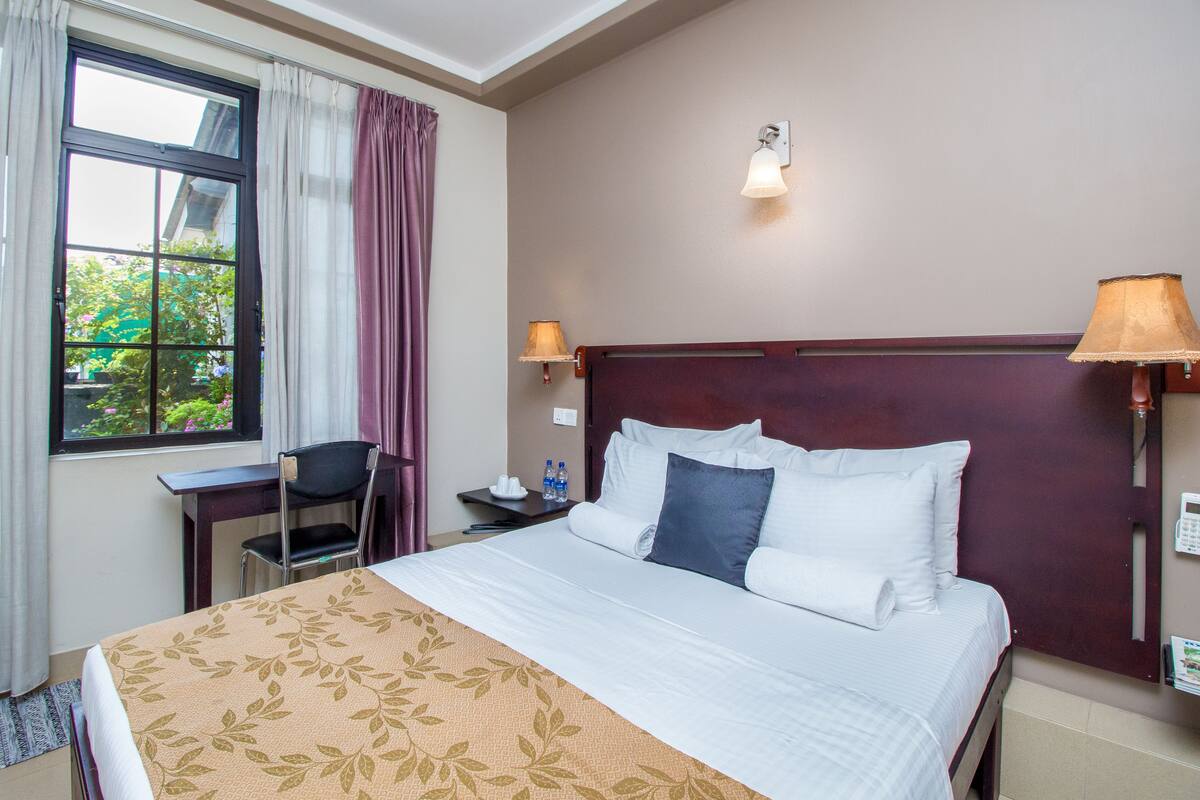 A hotel room with a bed, a desk, a chair, a window with curtains, and a wall-mounted lamp.