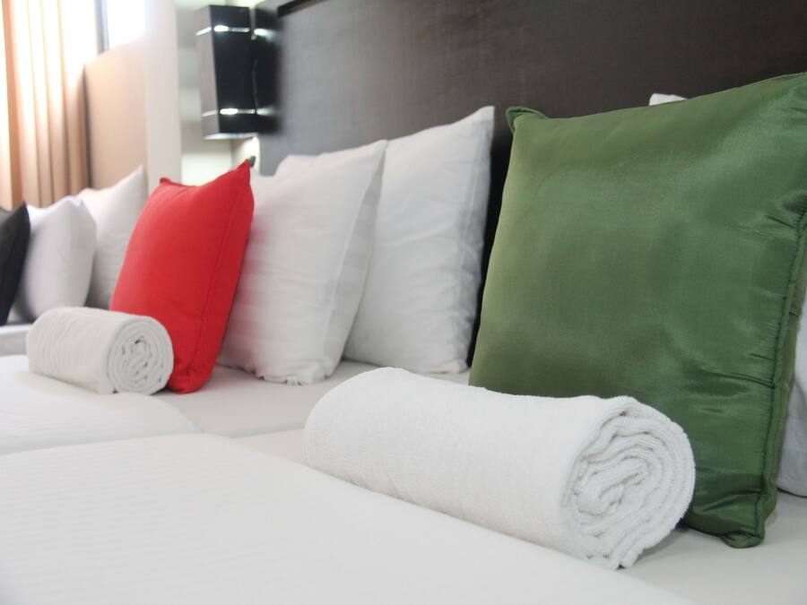 Una cama bien hecha con almohadas blancas y verdes, una toalla blanca enrollada y un cojín rojo.