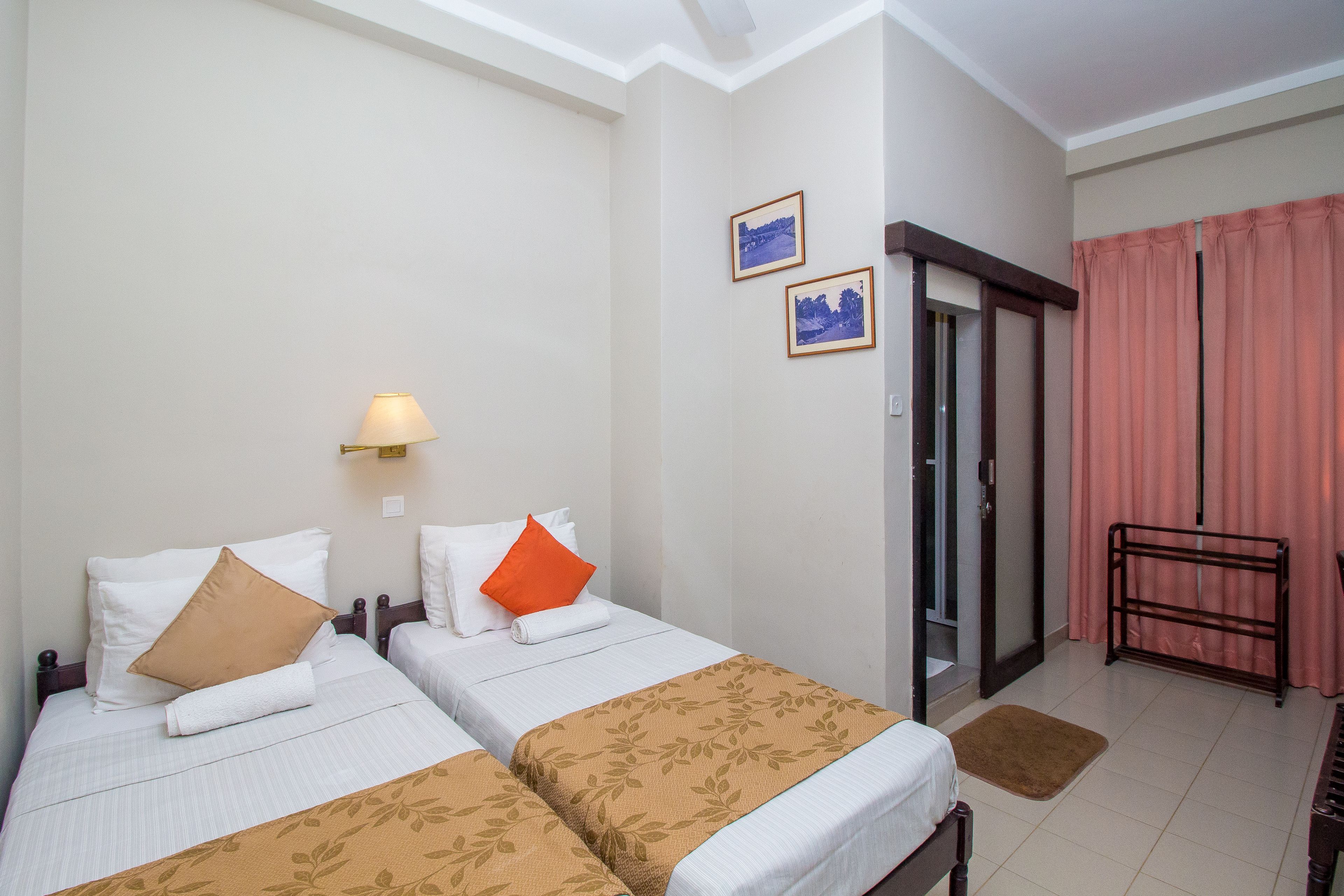 A hotel room with two beds, a nightstand, a lamp, a door with a mirror, and a window with curtains.
