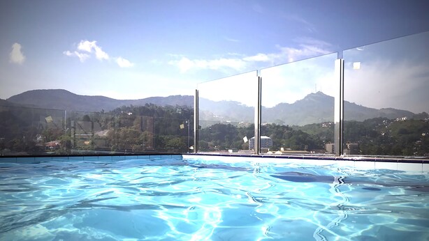 Ein Swimmingpool mit klarem, blauem Wasser, umgeben von Glasgeländern, mit Blick auf eine gebirgige Landschaft und Gebäude in der Ferne.