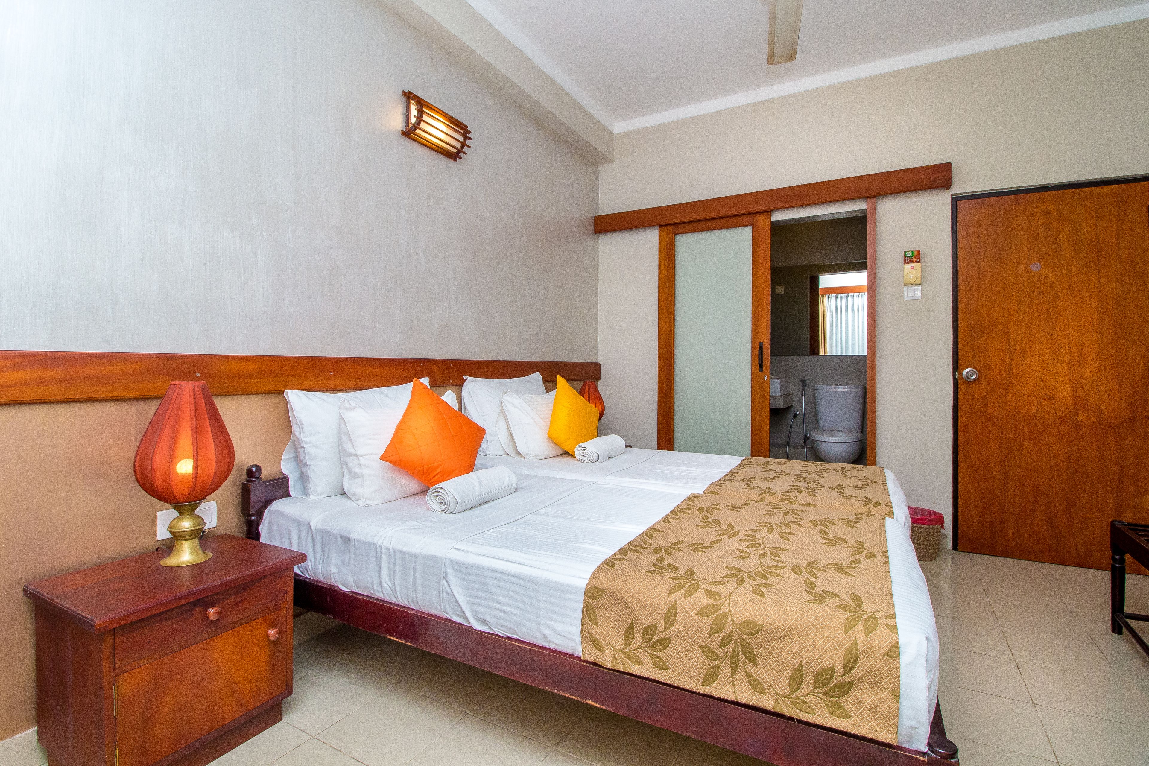 Una habitación de hotel con cama, mesita de noche, lámpara, cabecera de madera, espejo y puerta.