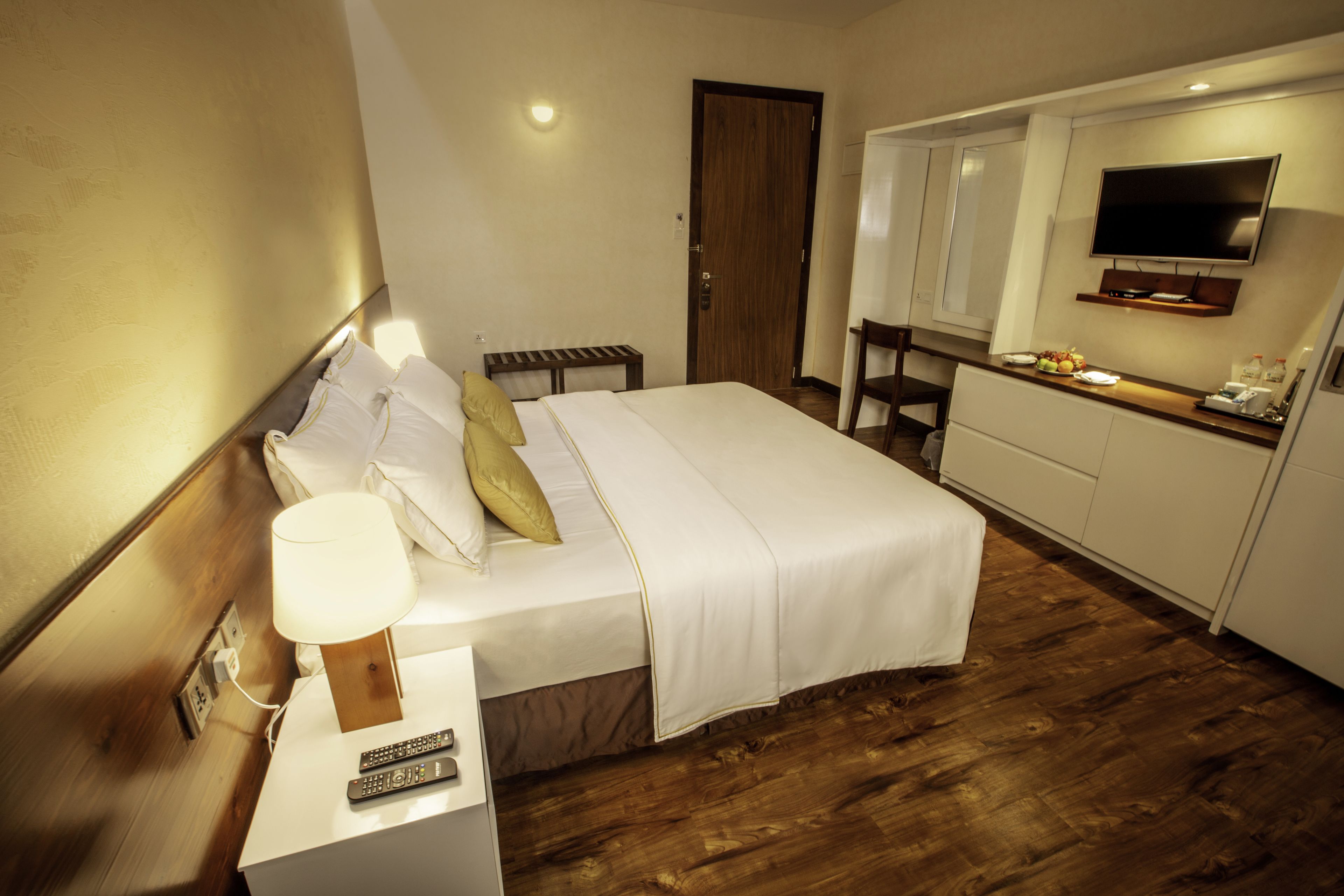 A hotel room with a bed, bedside table with a lamp, a TV mounted above a kitchen counter, and a wooden floor.