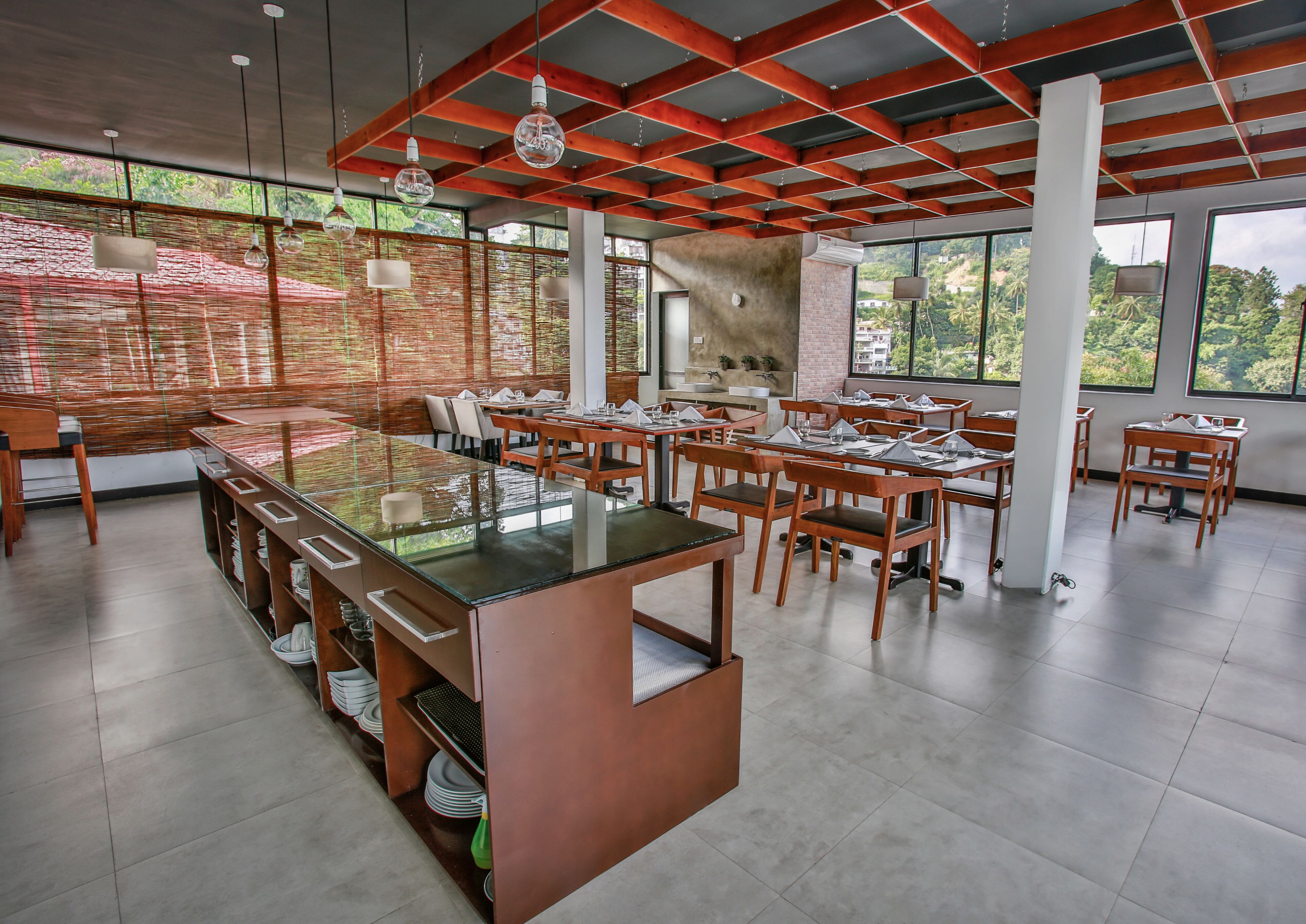 Een modern restaurant met een glazen eetgedeelte, houten tafels en stoelen, en grote ramen met uitzicht op groen.