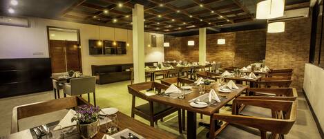 A restaurant with multiple dining tables and chairs, wooden beams on the ceiling, and pendant lighting.