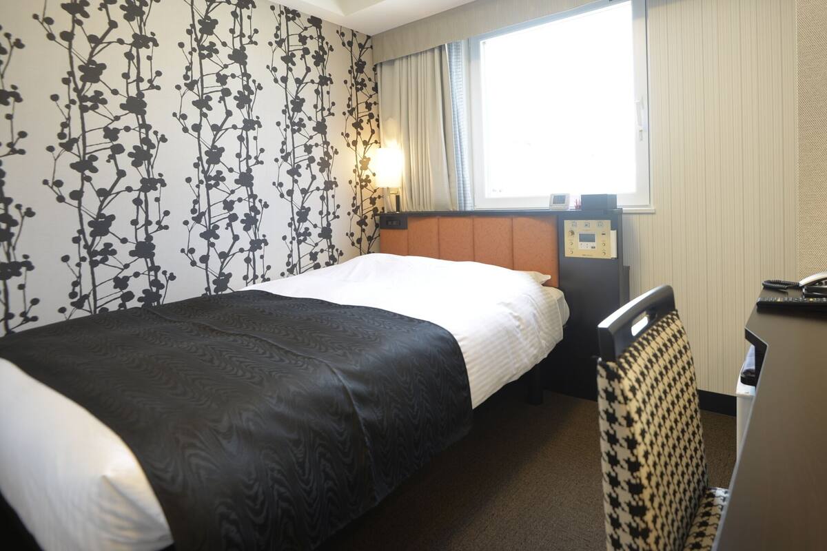 A hotel room with a bed, a desk with a telephone and a coffee maker, a window with curtains, and a wall with a floral pattern.