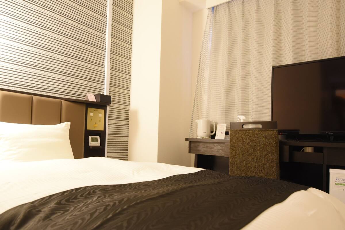 A hotel room with a bed, a bedside table with a clock, a television, and a window with blinds.