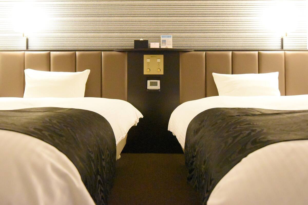 A hotel room with two beds, a headboard with a control panel, and a window with blinds.
