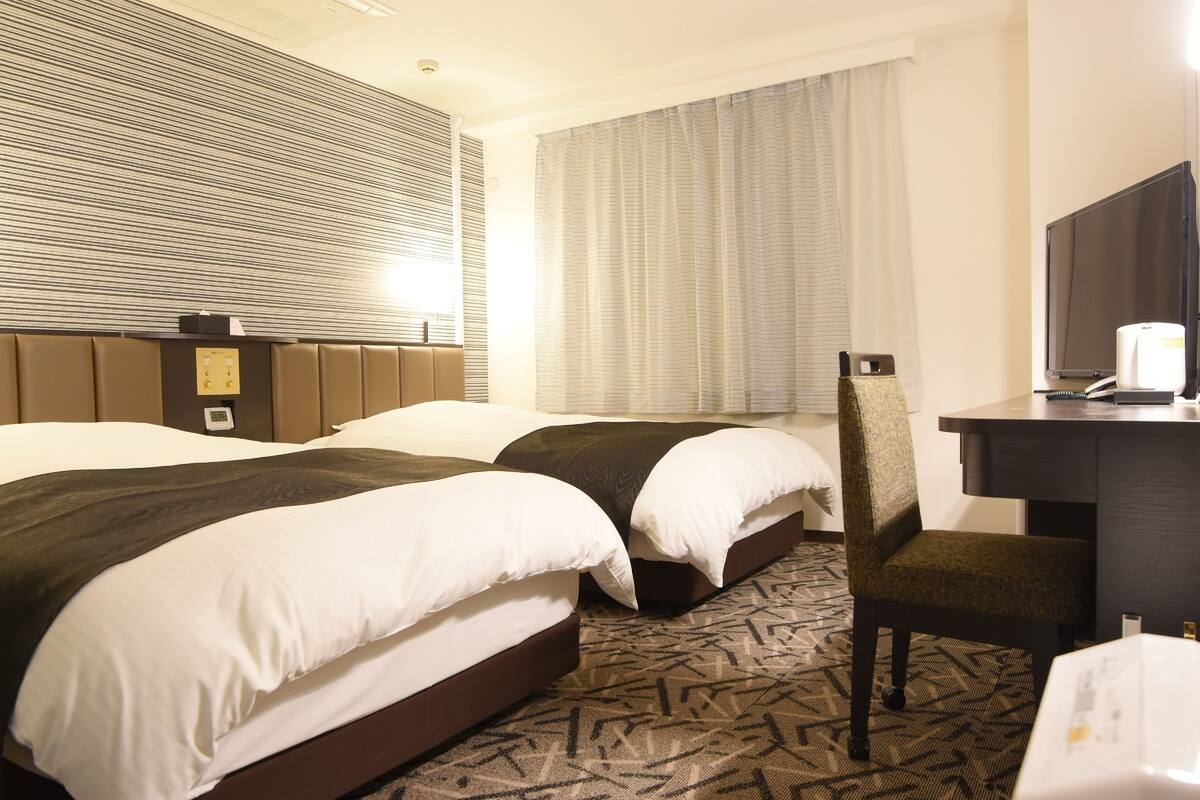 A hotel room with two beds, a desk, a chair, a television, and a window with curtains.