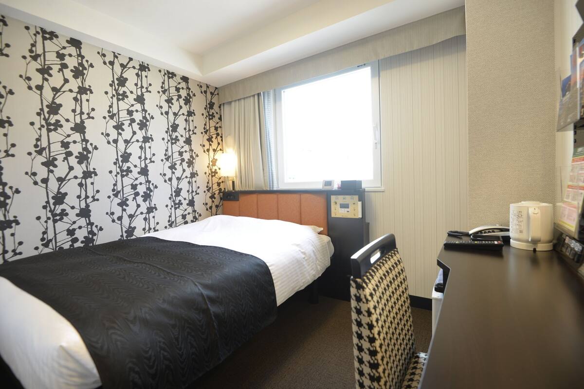 A hotel room with a bed, a desk with a chair, a window with curtains, and a wall with a floral pattern.