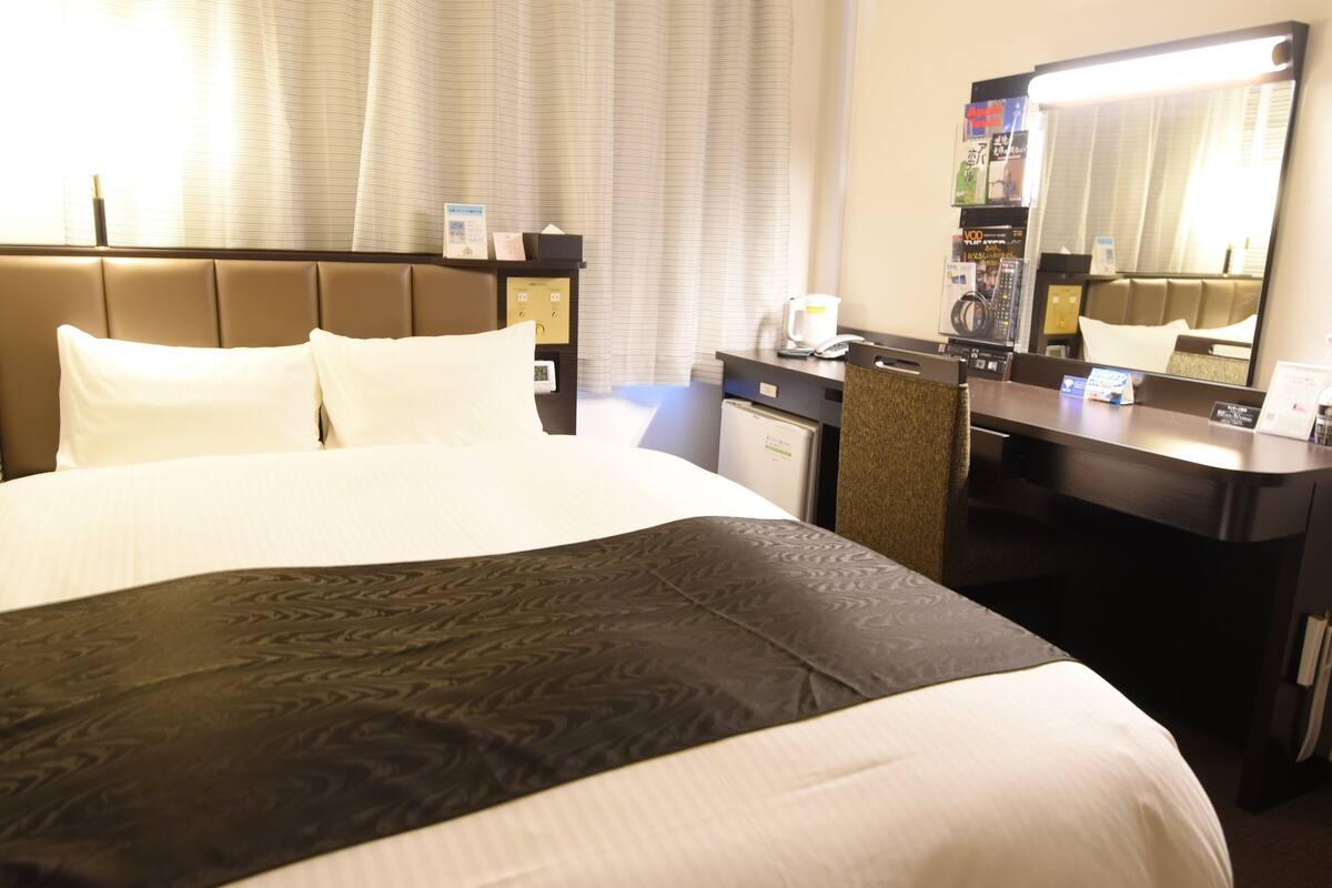 A hotel room with a large bed, a desk, a chair, a mirror, and a window with curtains.
