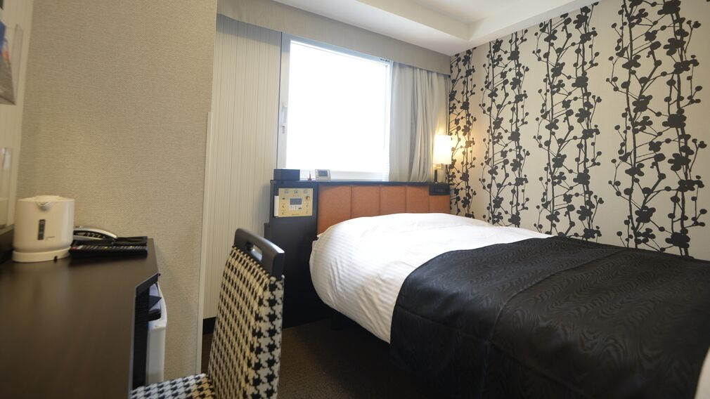 Una habitación de hotel con una cama, un escritorio con teléfono, una silla, una ventana con cortinas y una pared con estampado floral.