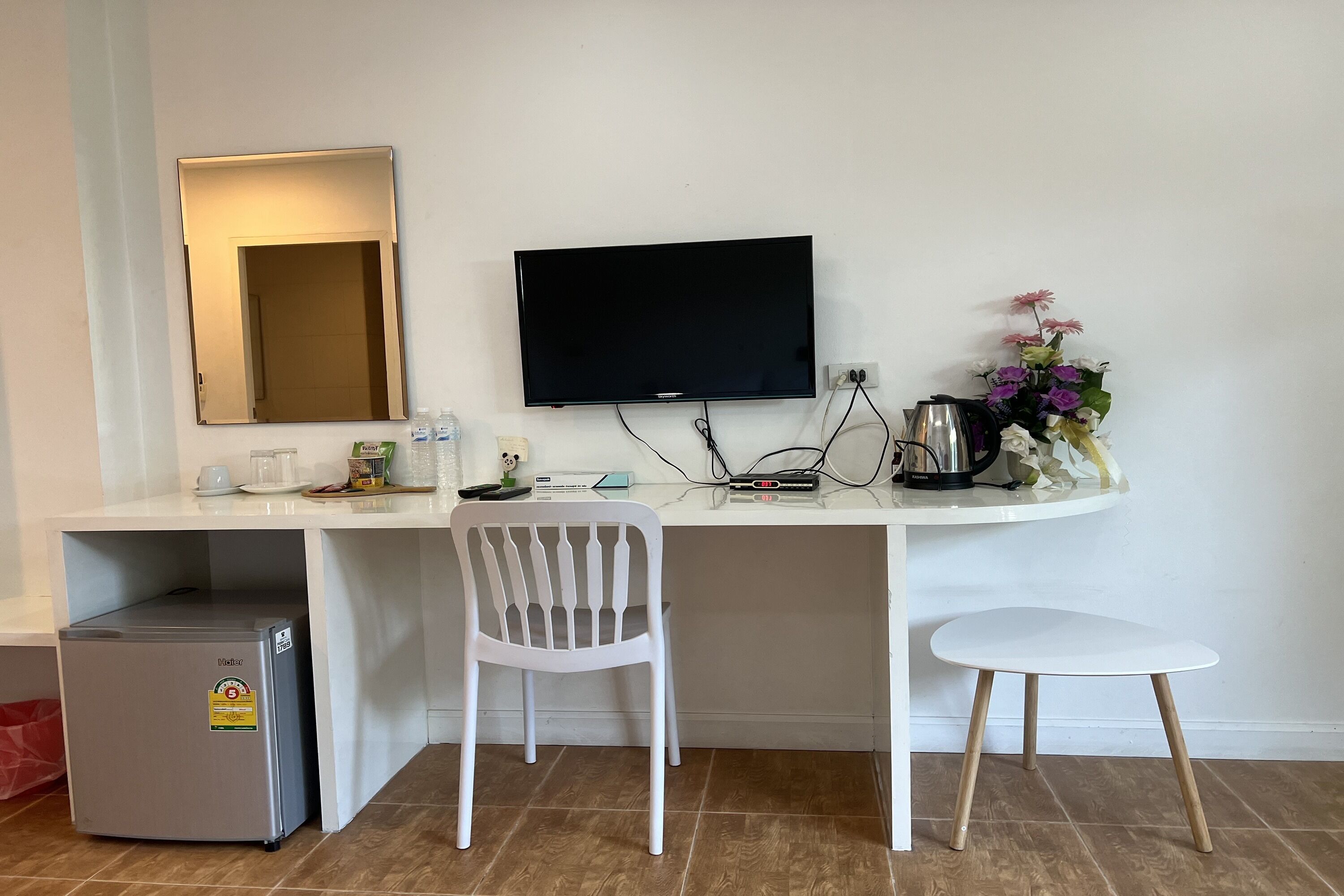 A compact hotel room with a small kitchenette, a desk with a flat-screen TV, a white chair, and a small refrigerator.