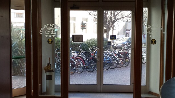 A hotel entrance with a glass door leading to a parking area with bicycles. - Hotel San Marco (Pesaro)