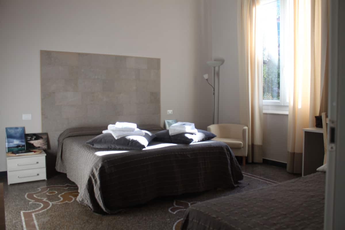 Un dormitorio con cama, mesita de noche, silla, lámpara de pie y ventana con cortinas.