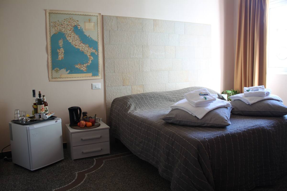 Habitación de hotel con cama, un escritorio pequeño con cafetera, un mapa en la pared y ventana con cortinas.