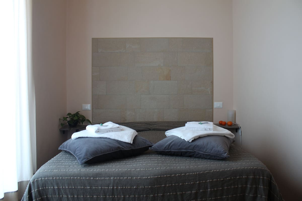 Habitación de hotel con cama, almohadas, una planta y un estante en la pared con toallas y naranjas.
