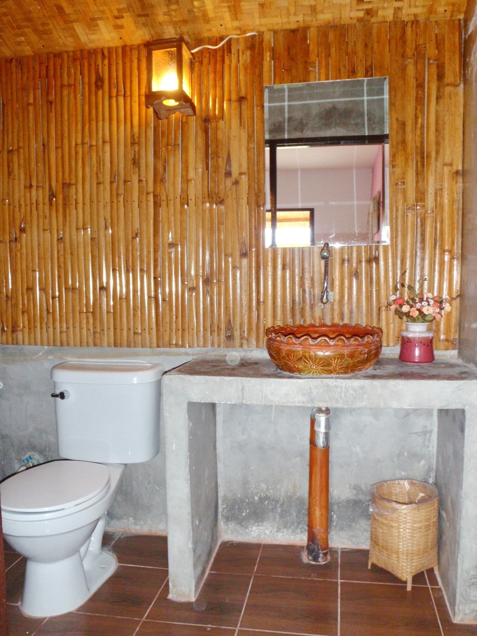 Une salle de bain avec un mur en bambou, des toilettes, un lavabo agrémenté d’un récipient décoratif, un miroir et une petite poubelle.