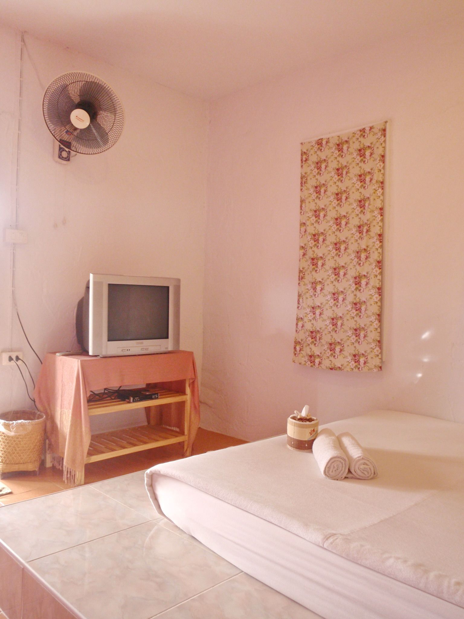A bedroom with a bed, a television on a wooden stand, a fan, and a wall tapestry.