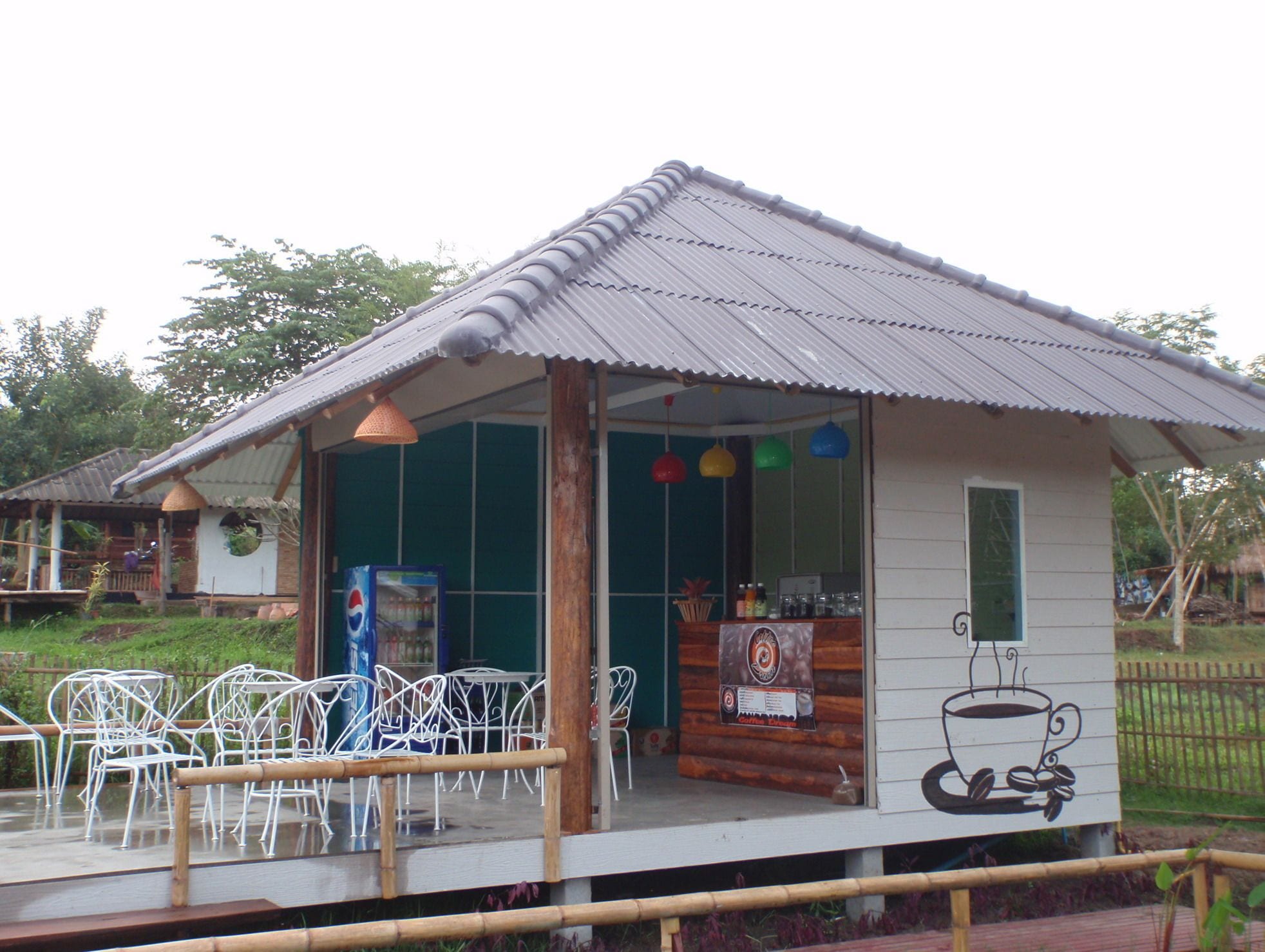 A small building with a corrugated metal roof, a glass door, and a sign featuring a coffee cup. There are white metal chairs outside.