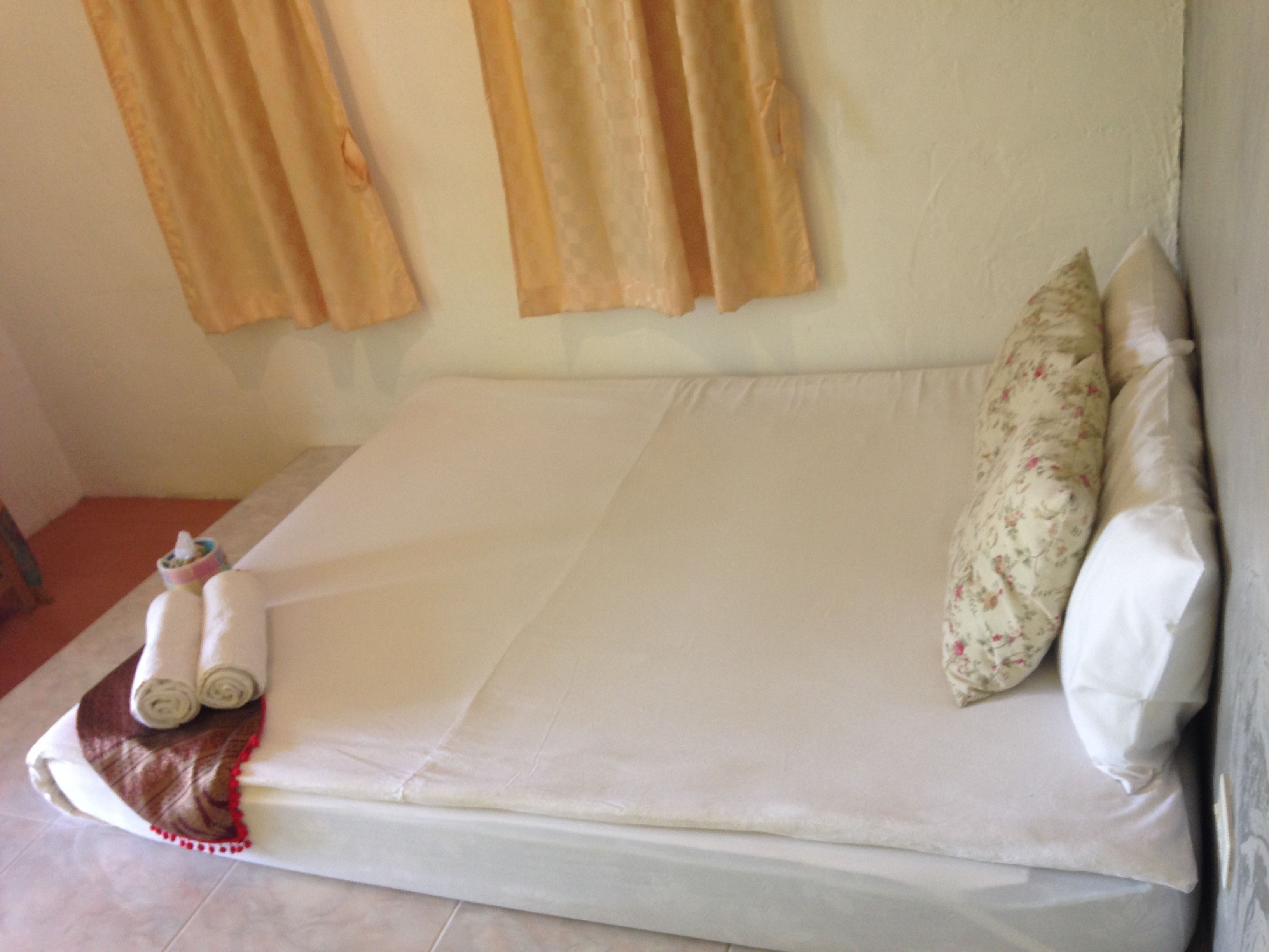 A neatly made bed with white linens, a floral pillow, and rolled towels on a white pillow.
