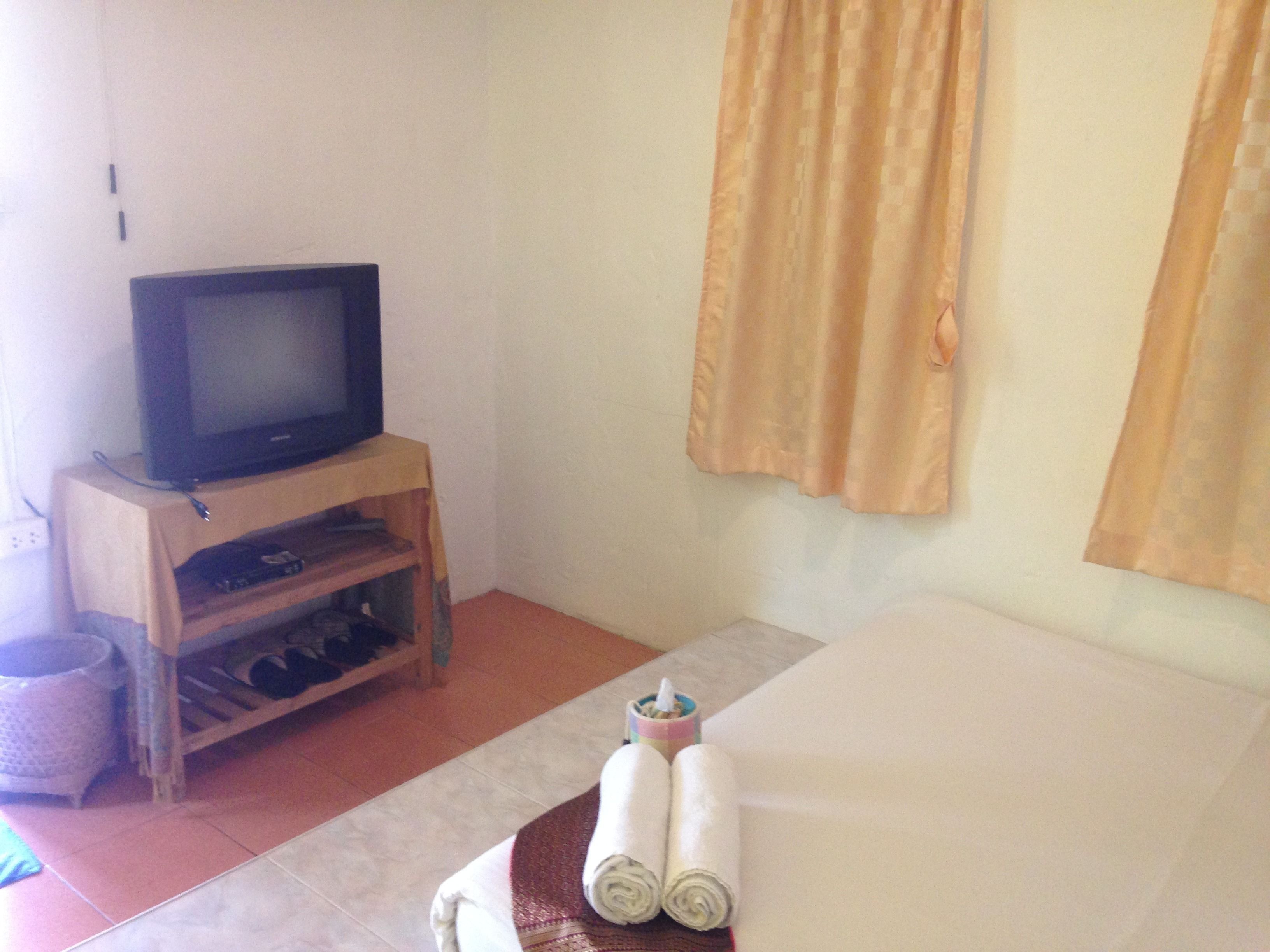 A hotel room with a television on a wooden stand, a bed with rolled towels, and a curtain on the window.