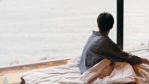 Iemand zit op een bed en kijkt uit het raam op zee.