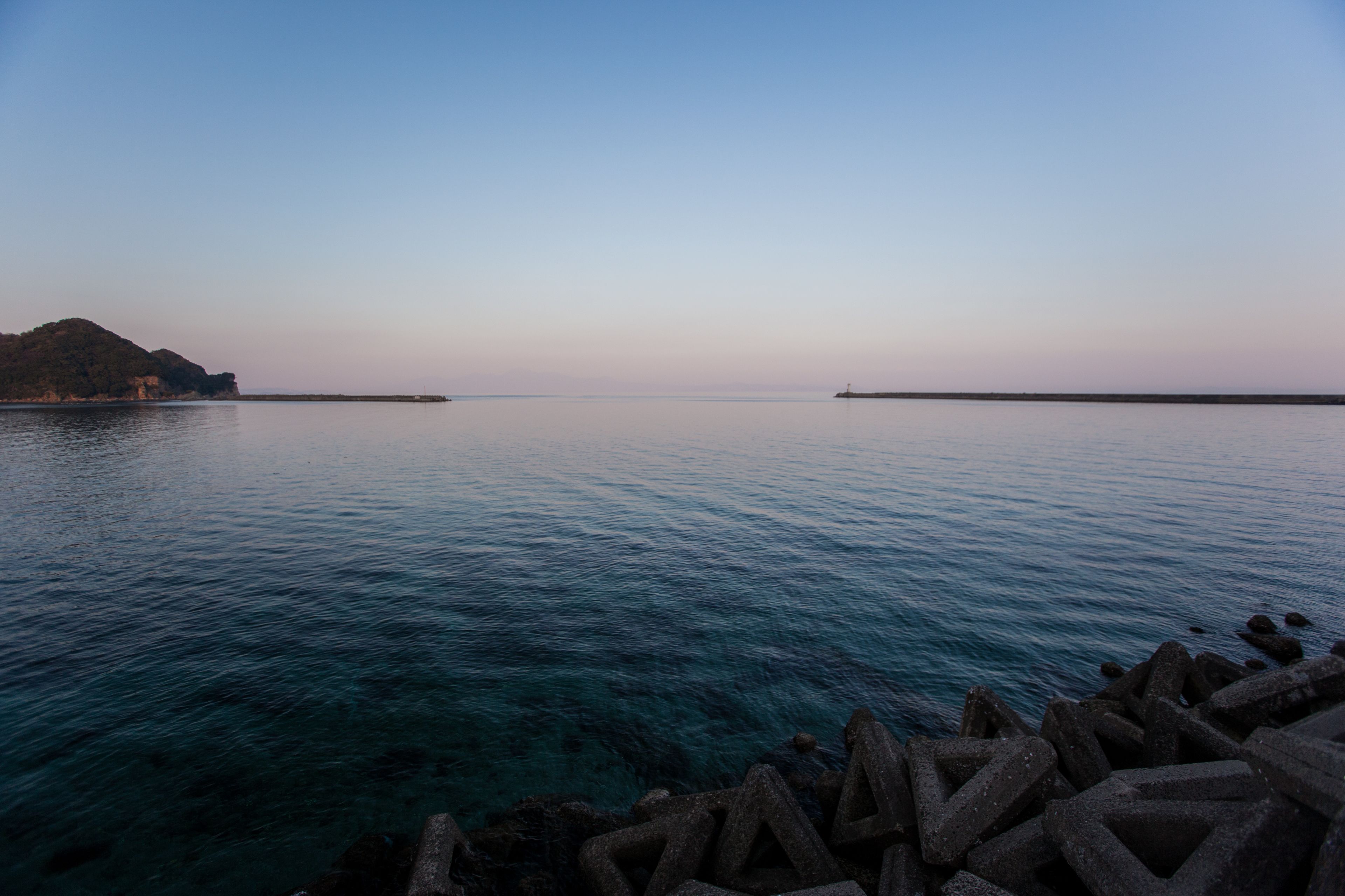Mar en calma con aguas cristalinas, un rompeolas rocoso y una isla en la distancia.