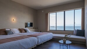 Een slaapkamer met een groot bed, een aan de muur bevestigde televisie, een raam met uitzicht op zee en een klein tafeltje met een vaas.