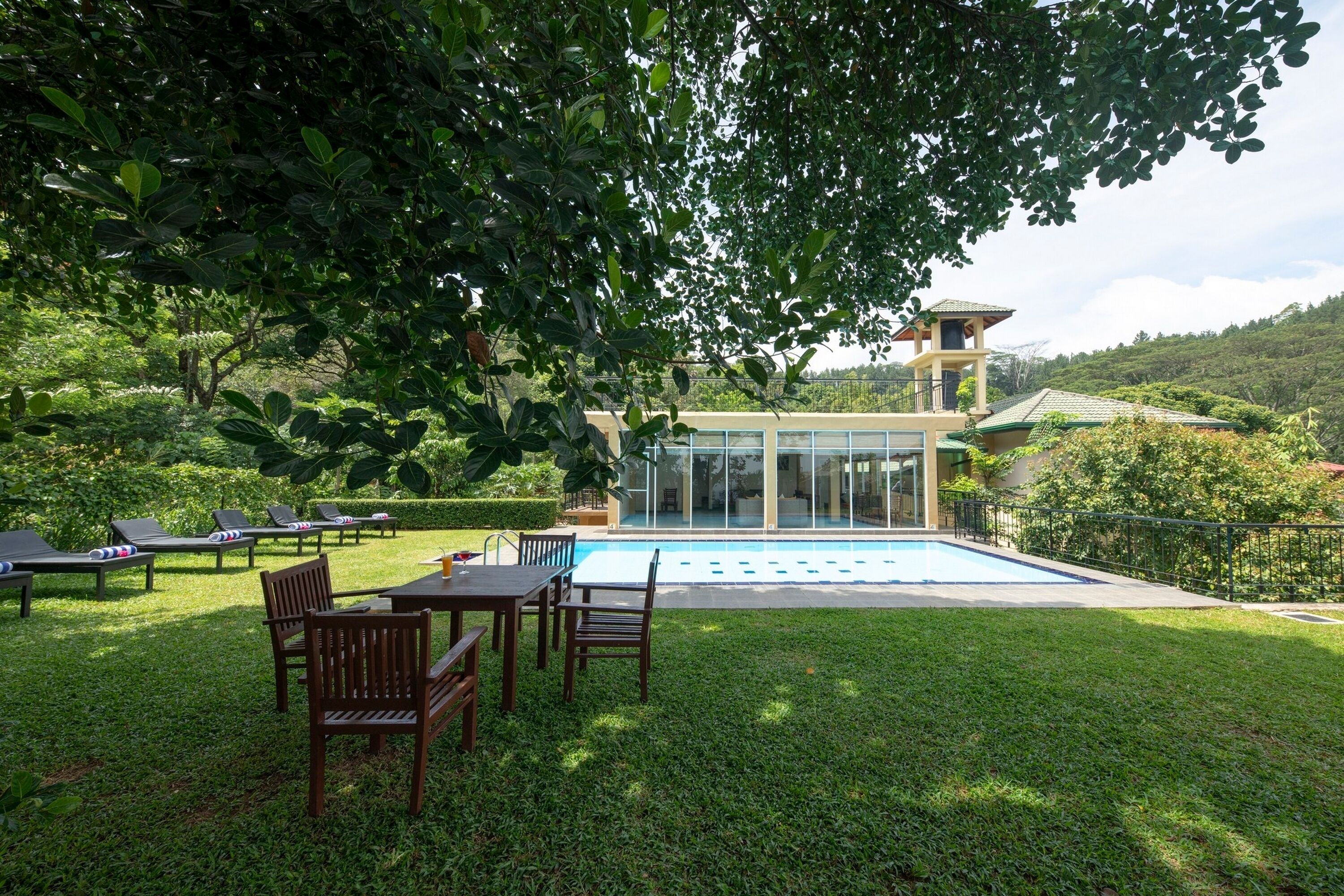 A pool area with lounge chairs and a dining table, surrounded by lush greenery and a modern building with large windows.