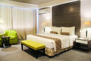 Habitación de hotel con una cama grande, un sillón verde, una mesita, un banco y dos mesitas de noche con lámparas.