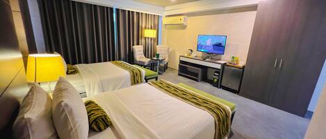 Quarto de hotel com duas camas, uma escrivaninha, uma cadeira, uma televisão e um guarda-roupa.