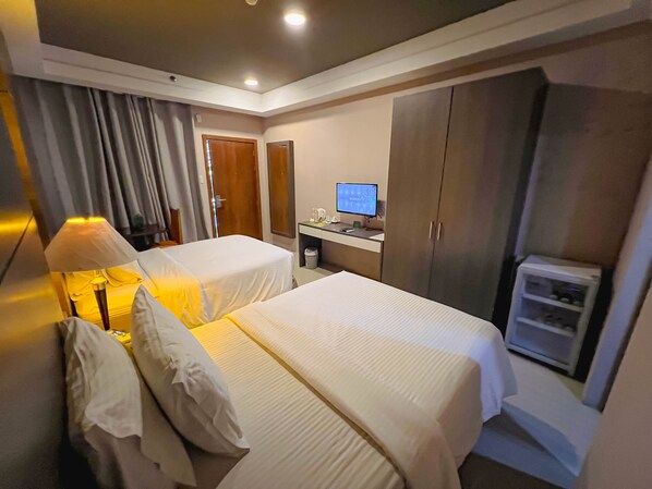 A hotel room with two beds, a desk with a computer, a wardrobe, and a small air conditioning unit.
