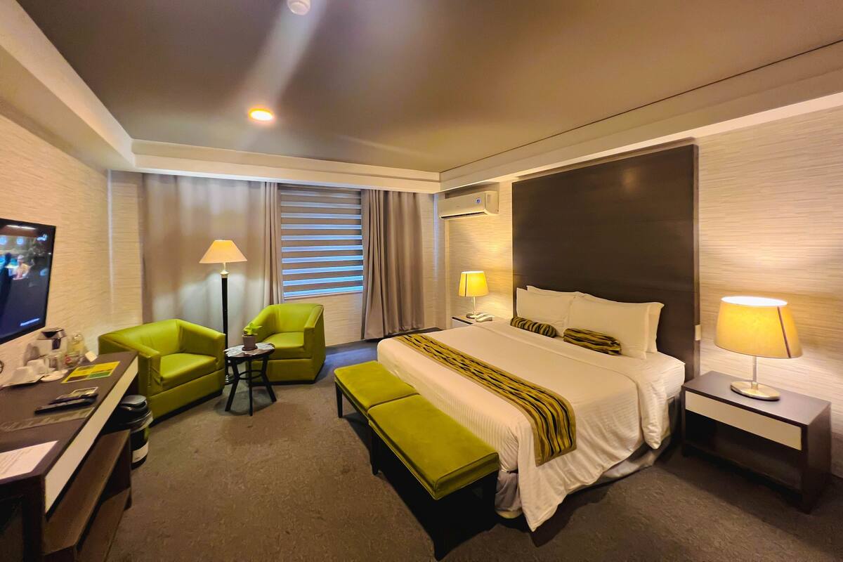 A hotel room with a bed, a desk, a chair, a bench, and a TV.