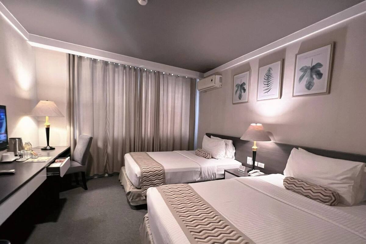 A hotel room with two beds, a desk, a chair, a TV, and a window with curtains.