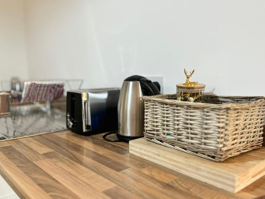 Une table en bois sur laquelle se trouvent un appareil électronique noir, une bouilloire en acier inoxydable et un panier en osier contenant un objet décoratif.